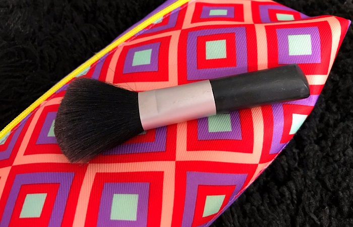 Read on for tips on how to clean makeup brushes. Pictured is a black makeup brush against a bright pink and purple background. 