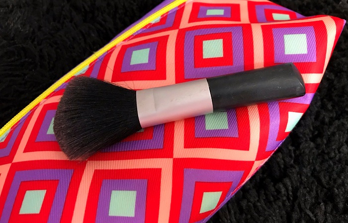 Read on for tips on how to clean makeup brushes. Pictured is a black makeup brush against a bright pink and purple background.