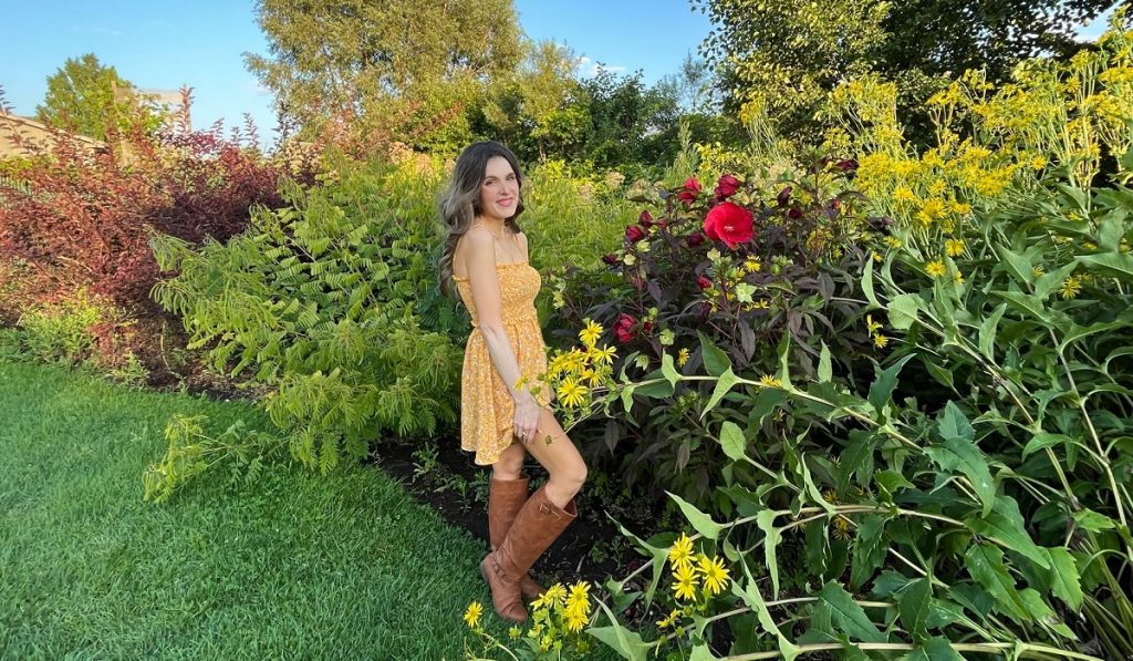 Image of Anne Erickson from WRIF, Audio Ink Radio and Upon Wings, standing at the MSU gardens on a sunny day. She is surrounded by flowers and blue skies.