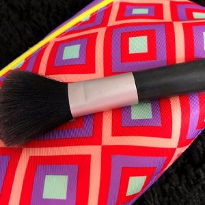 Read on for tips on how to clean makeup brushes. Pictured is a black makeup brush against a bright pink and purple background.
